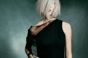 Person with short, platinum blonde hair covering part of their face, wearing a sleeveless black dress with a sheer shoulder detail, stands against a dark background—embodying modern style inspired by wellness rituals.