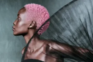 A person with short pink hair, vibrant from aveda hair care, poses in profile against a gray background, wearing a sheer black garment that fans out dramatically beside them.