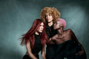 Three women pose together against a muted background, each with distinct hairstyles—long red hair, curly blonde hair, and short pink hair—showcasing the results of botanical hair treatment. All wear black outfits.
