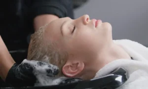 A person with closed eyes lies back as their hair is washed with shampoo at Thirty Hair salon, a soothing moment of self-care rituals, wrapped in a white towel around their neck.