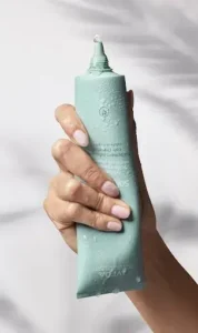 A hand holding a teal-colored tube of Aveda product with some moisture drops visible on the surface, perfect for achieving radiant hair with demi color shine.