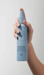 A hand holding a blue bottle of Aveda Smooth Infusion Perfect Blow Dry spray, perfect for maintaining enhanced existing color, against a plain light background.
