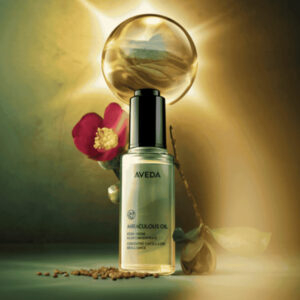 A bottle of Aveda Miraculous Oil, perfect for scalp care and a hair reset, is displayed with a pink flower, golden seeds, and a glowing orb in the background.