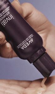 A person dispenses cream from a dark purple Aveda tube onto their open palm.