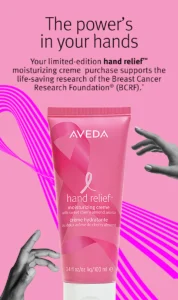 Pink tube of Aveda hand relief moisturizing creme with breast cancer ribbon, text about supporting Breast Cancer Research Foundation, and a hand reaching toward the tube.