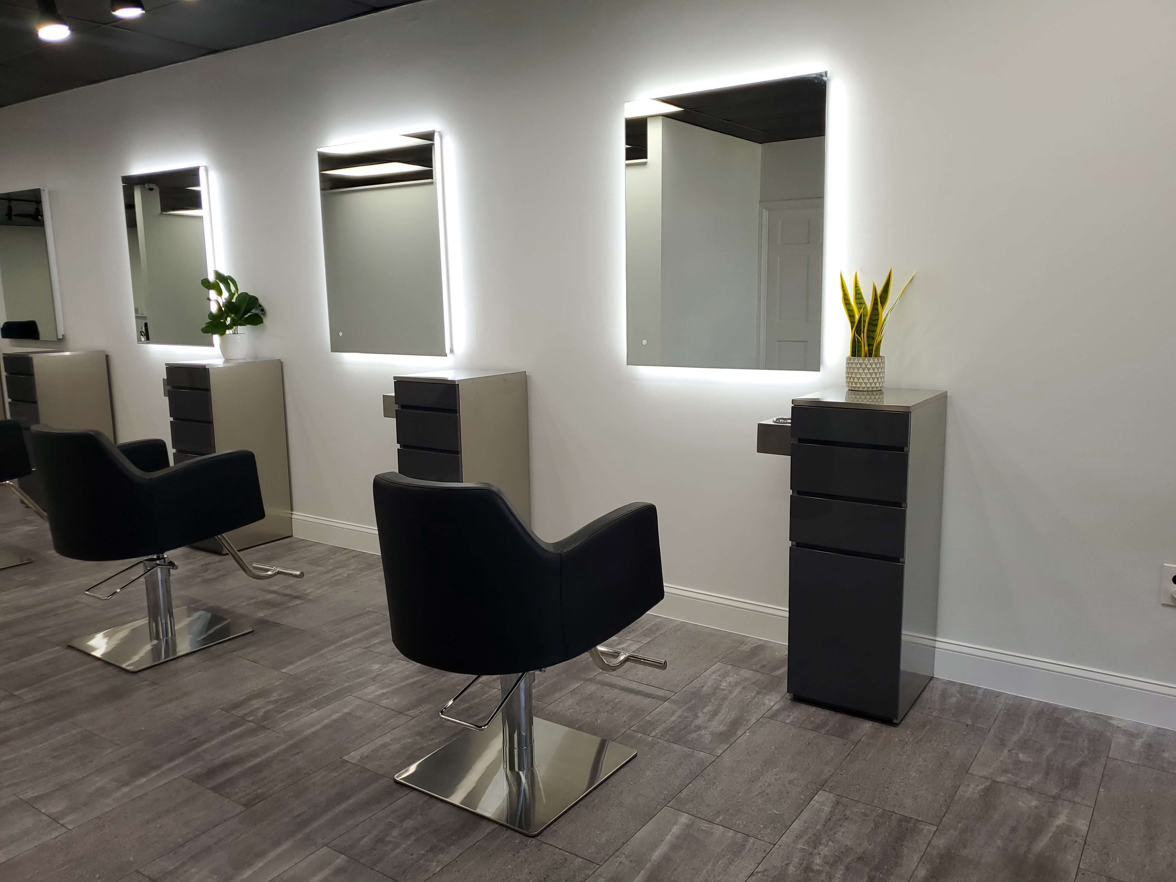 A modern salon interior with black styling chairs, large illuminated mirrors, gray cabinets, and potted plants on top of the cabinets against a white wall.