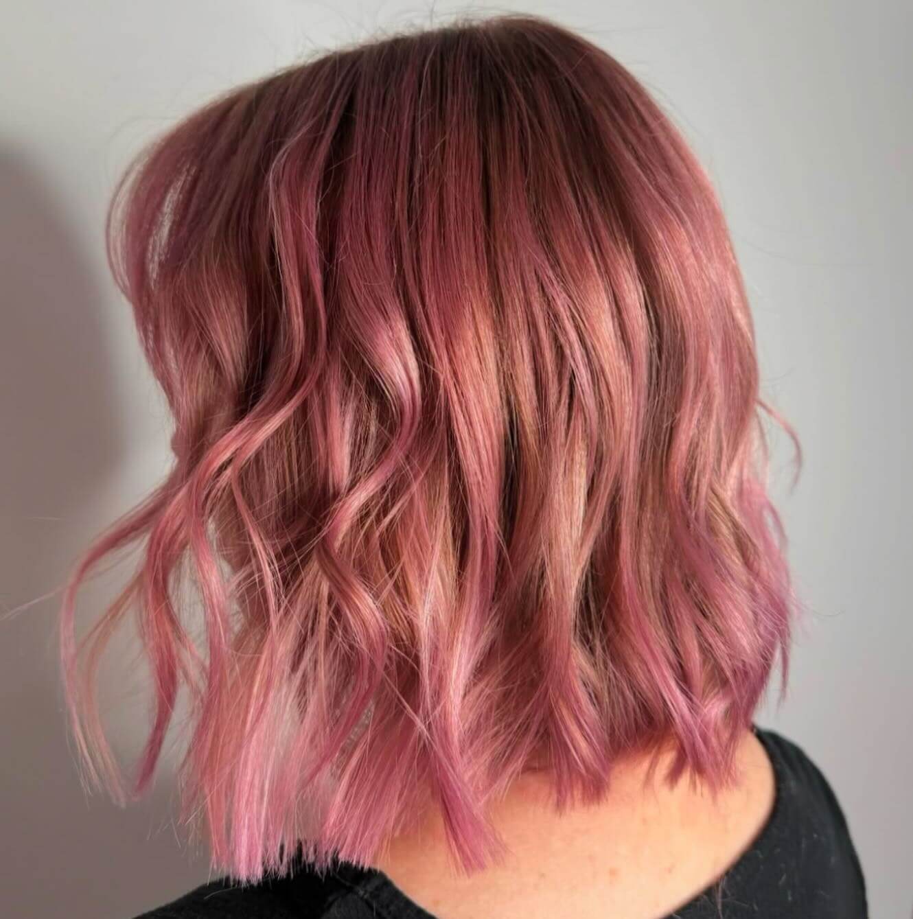 Person with short, wavy hair dyed in shades of pink and purple, photographed from behind against a plain background.