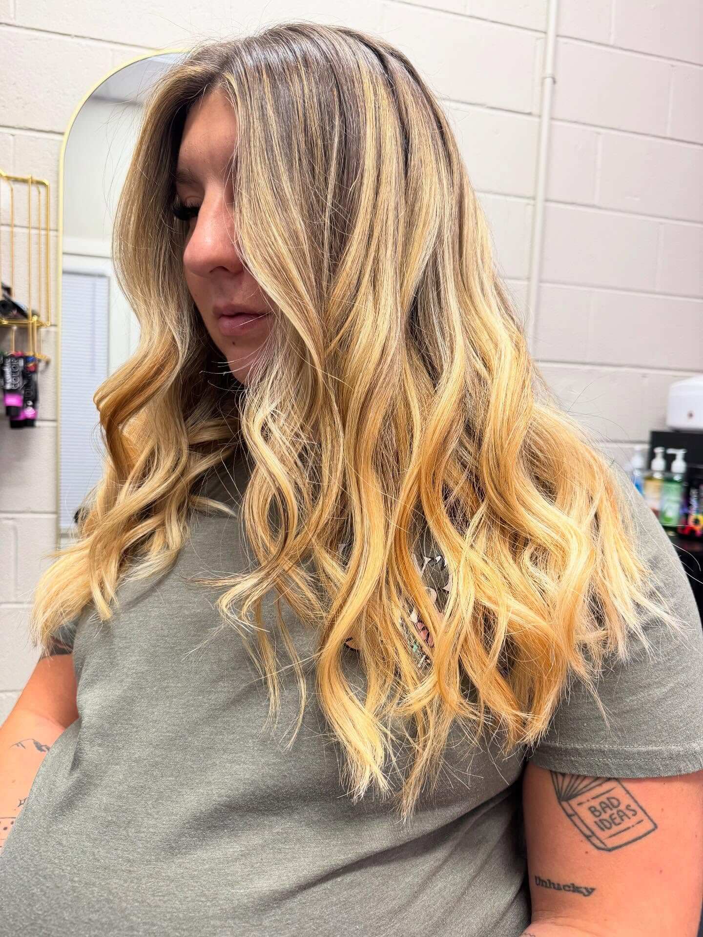 Woman with long, wavy blonde hair featuring darker roots stands indoors, wearing a gray shirt; tattoos visible on her arm.