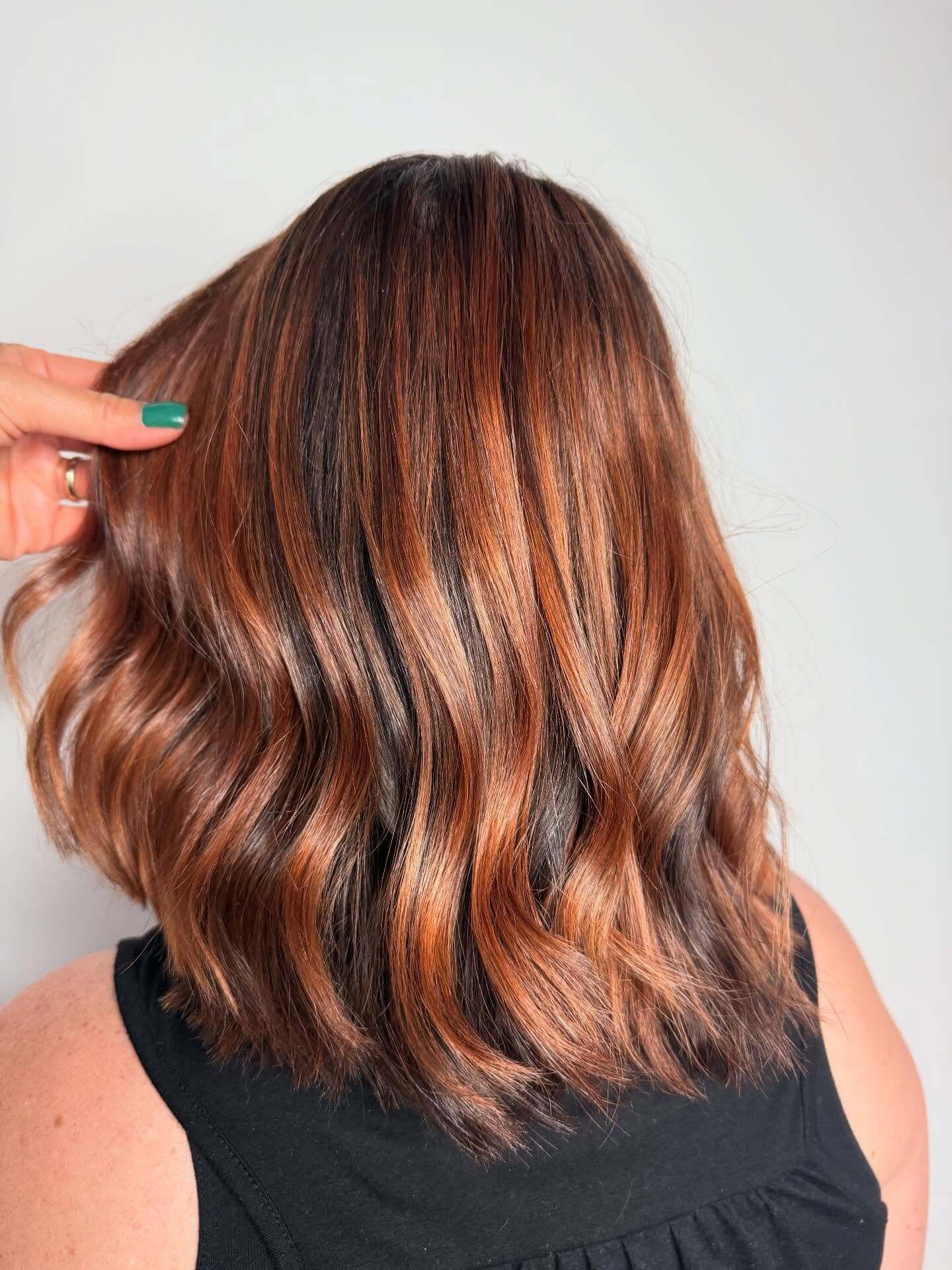 Person with shoulder-length, wavy hair featuring a blend of copper and dark brown tones, wearing a black sleeveless top. A hand with green nail polish is touching the hair.