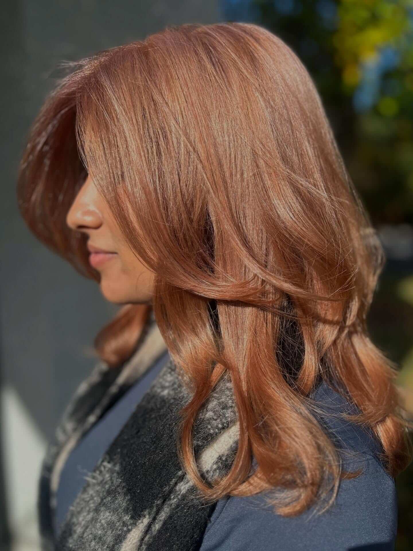 A woman with long, layered, rose gold hair is shown in profile, standing outdoors in natural light.