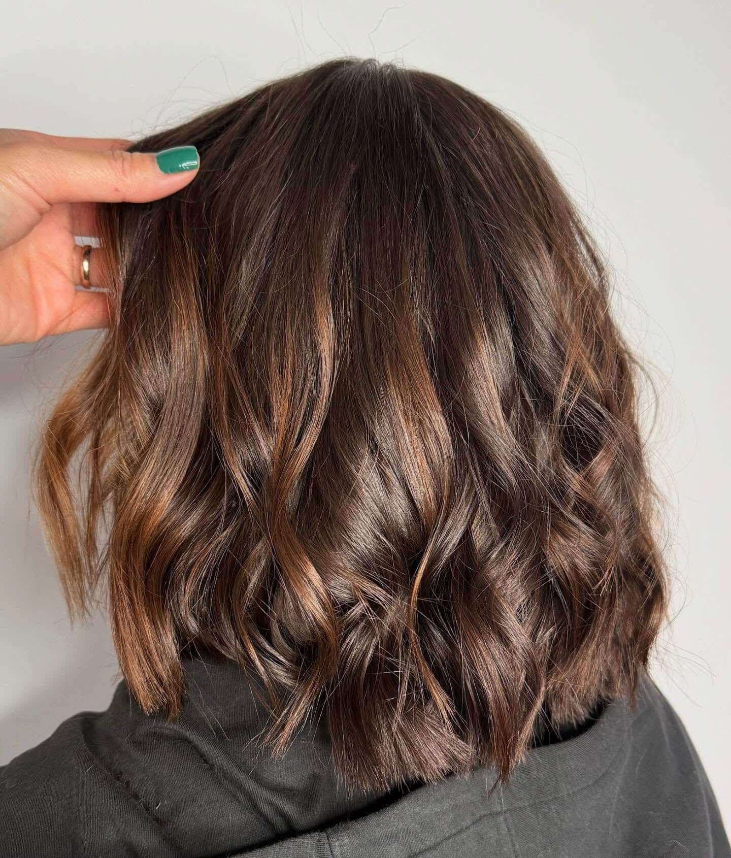 A hand with green nail polish holds the side of a person's head, showing shoulder-length, wavy brown hair with subtle highlights.