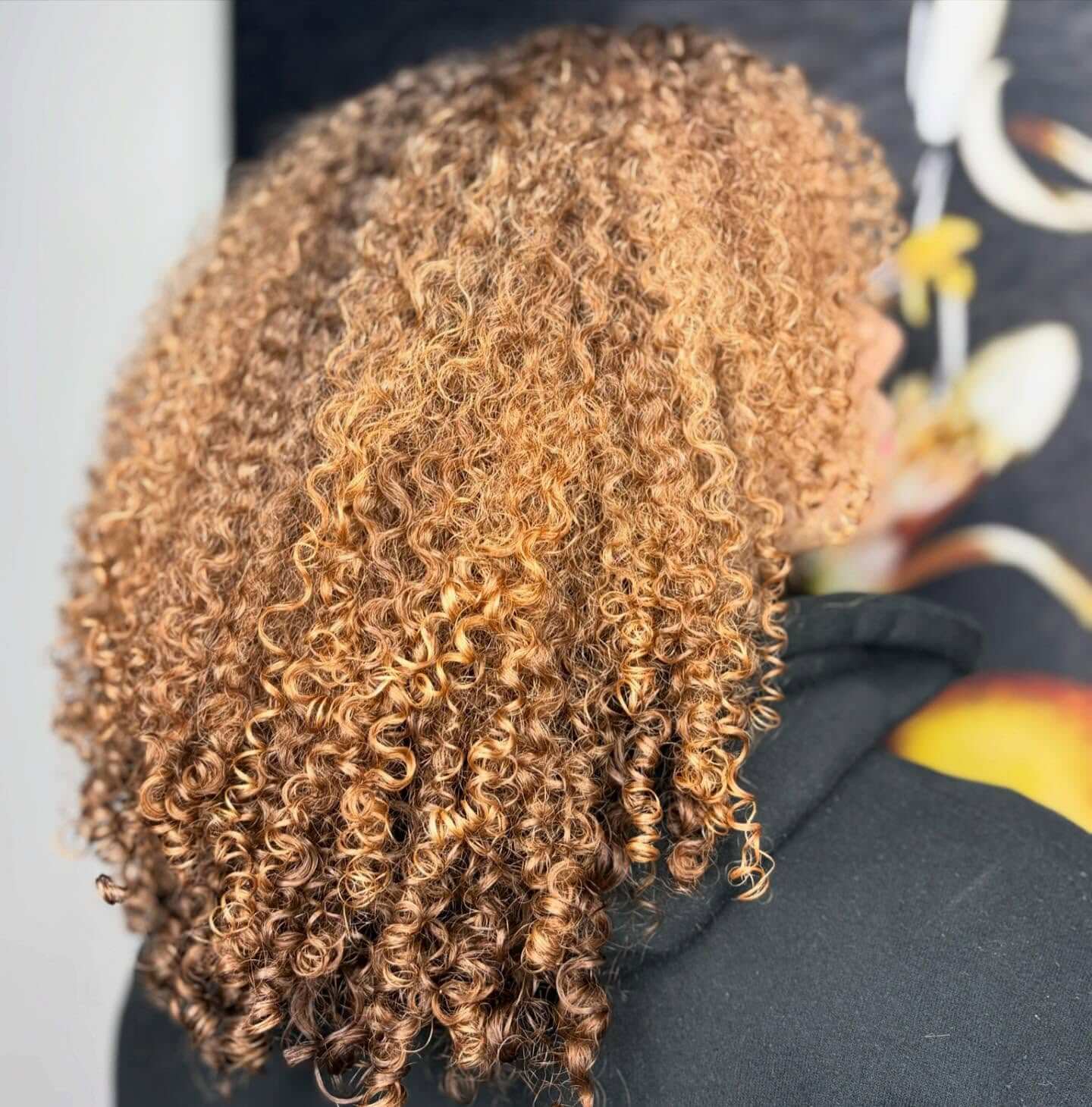 Person with shoulder-length, tightly coiled, light brown curly hair, photographed from behind against a dark, blurred background.