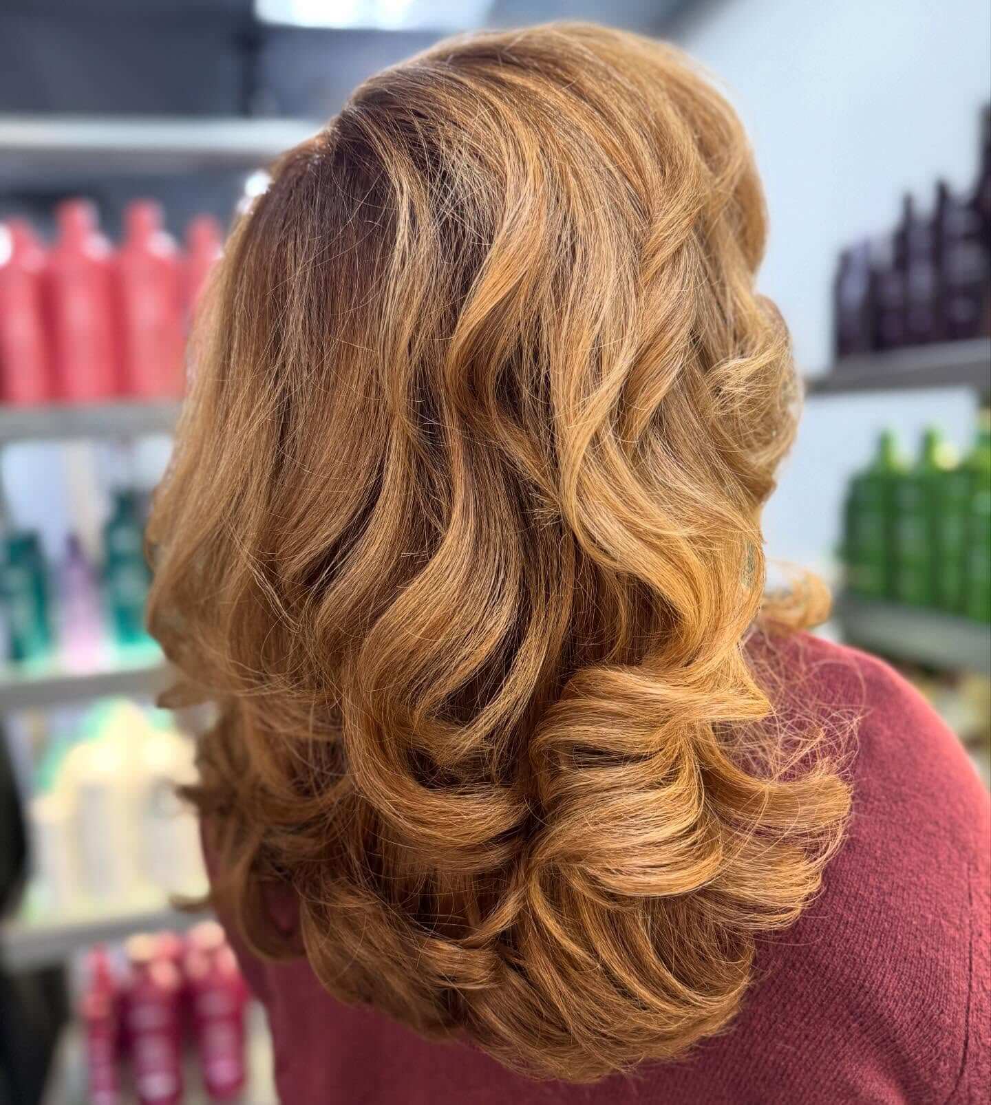 A person with shoulder-length, voluminous wavy hair in a light copper color, shown from the back, standing in front of shelves with hair products.