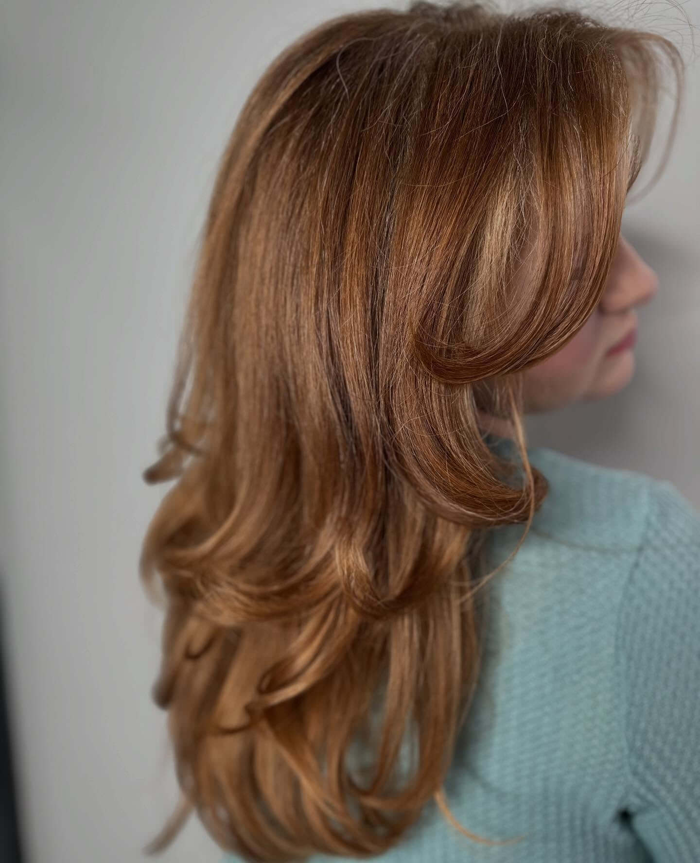 A person with long, layered, light brown hair styled in loose waves, wearing a textured light blue top, is turned slightly away from the camera.