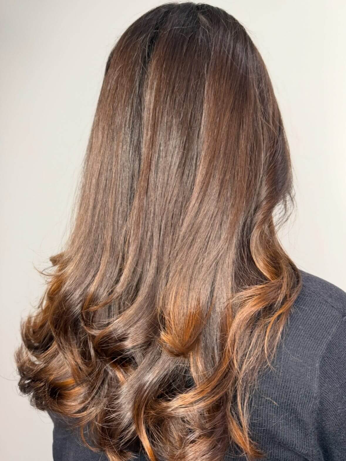 A person with long, smooth, brown hair featuring soft curls at the ends, photographed from behind against a plain background.