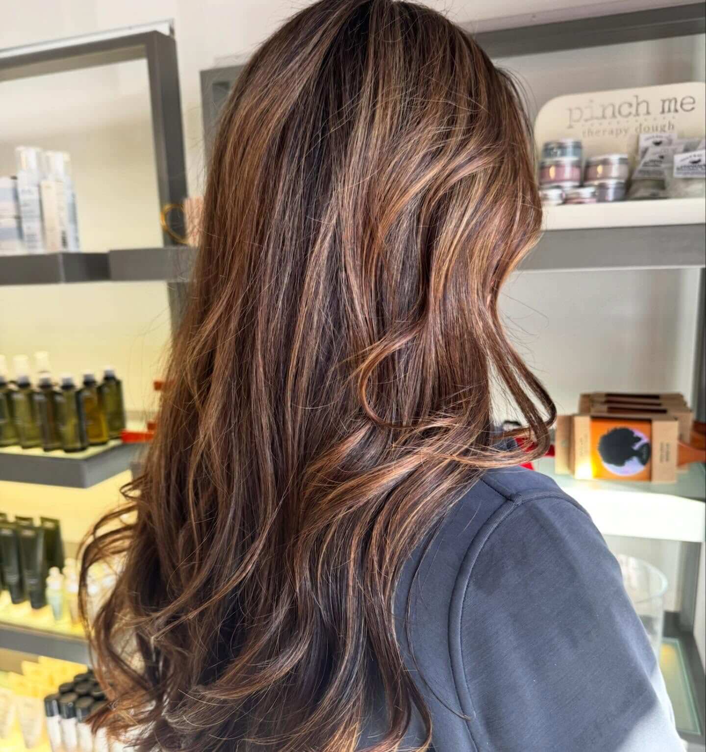 Person with long, wavy brown hair with caramel highlights, standing in front of shelves with hair and skincare products in a salon.