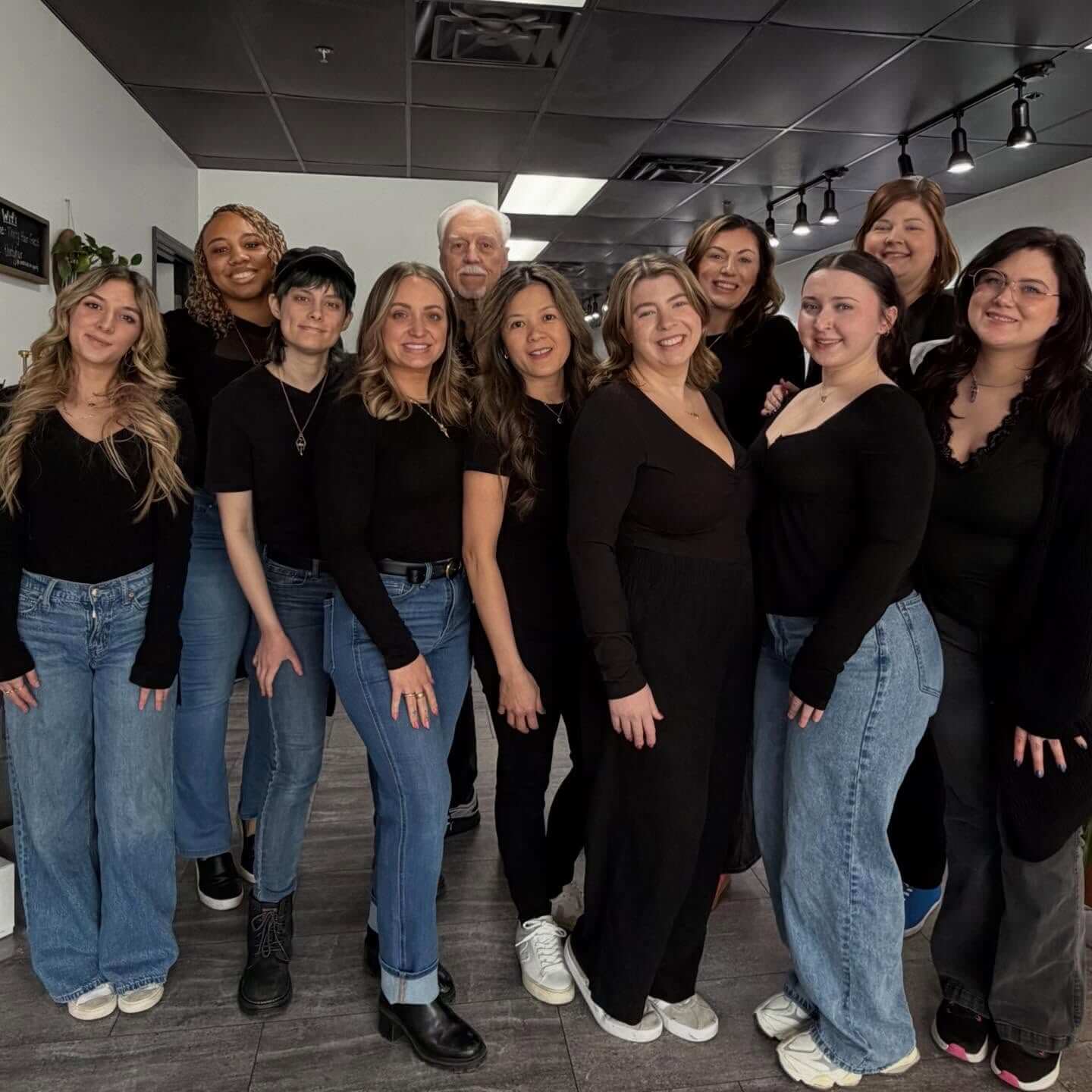 A group of eleven people pose indoors, all wearing black tops and blue jeans or pants, standing on a gray floor under a black ceiling.