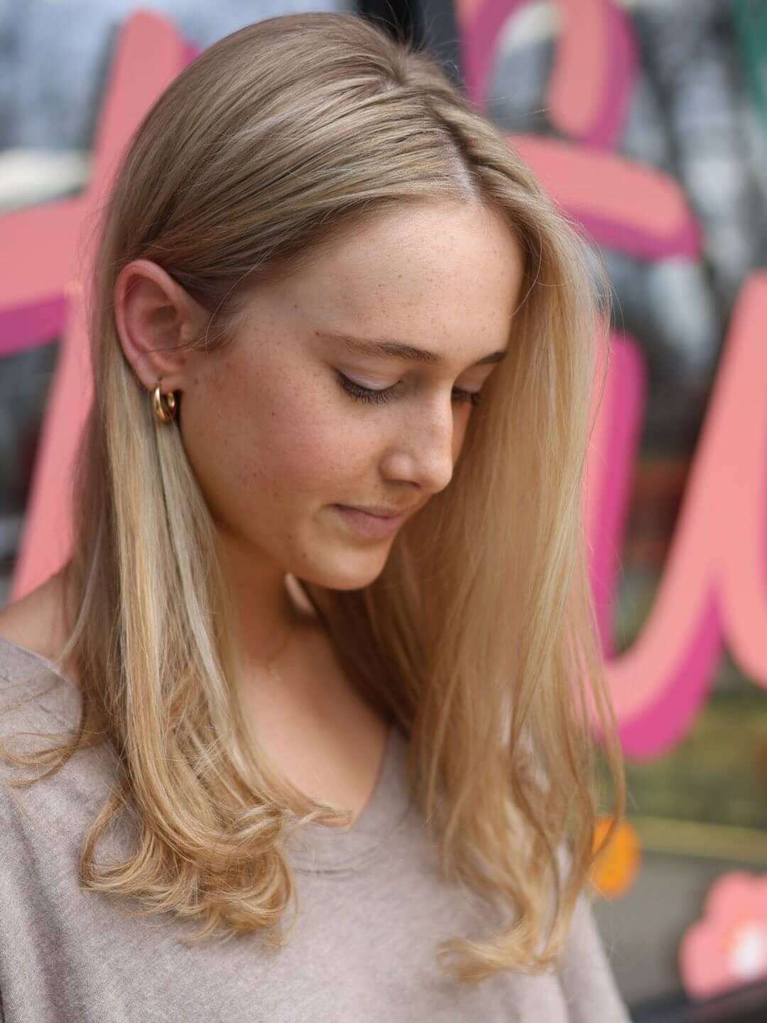 A woman with long blonde hair looks down, standing in front of a window with colorful shapes and letters in the background.