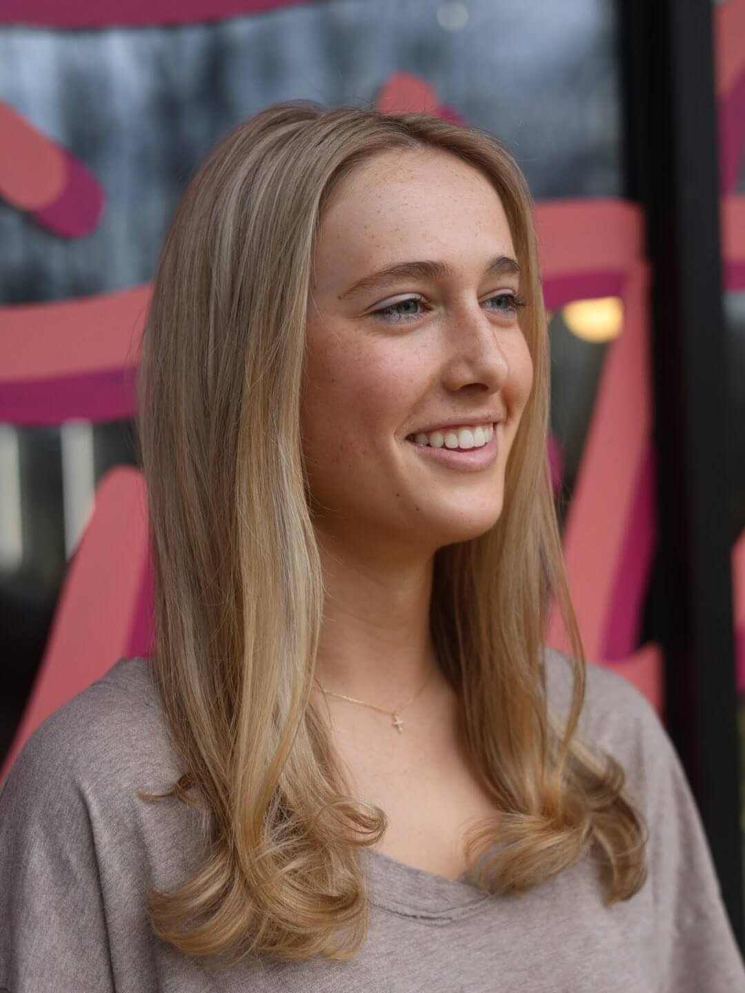 Young woman with long blonde hair and a light brown top stands indoors, smiling and looking to the side; abstract pink and red shapes are visible in the background.
