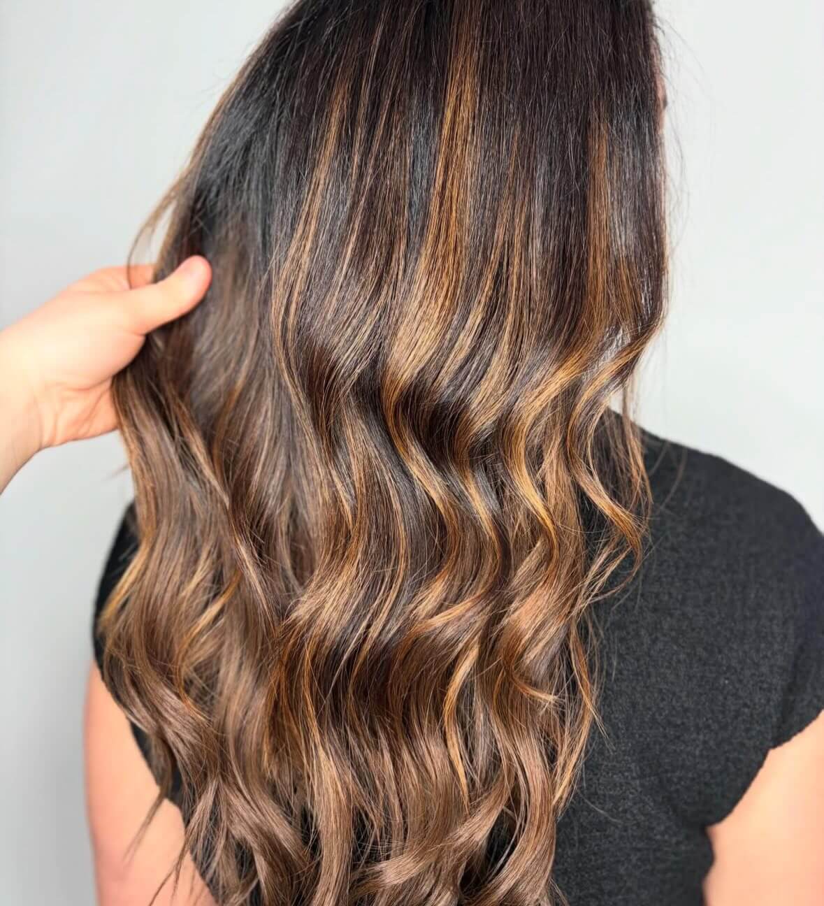 A person with long, wavy brown hair featuring caramel highlights is shown from the back; one hand is holding a section of the hair.