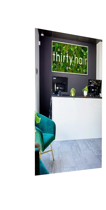 Reception desk with two computer monitors, a small potted plant, and a wall sign reading "thirty hair" on a green background. Green chair visible in the foreground.