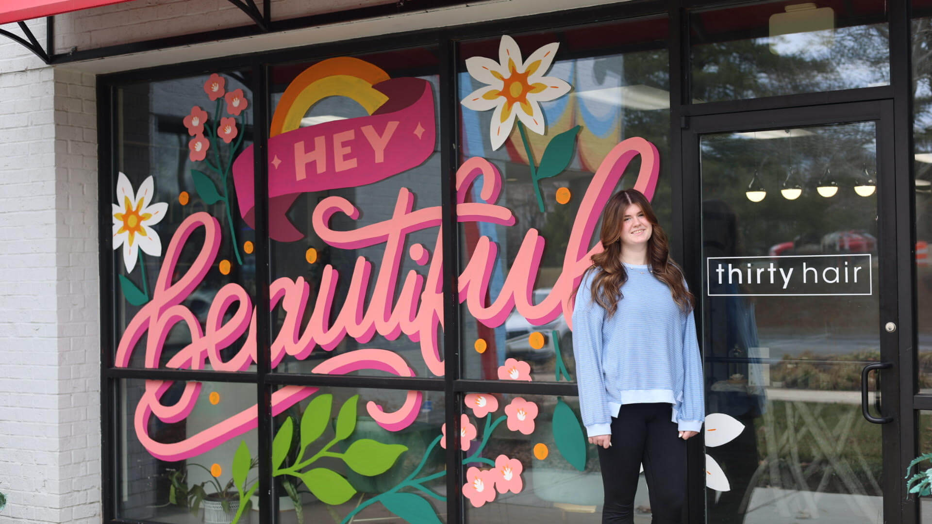 A person stands in front of a salon storefront with large colorful “Hey Beautiful” artwork on the window and a sign that reads “thirty hair.”.