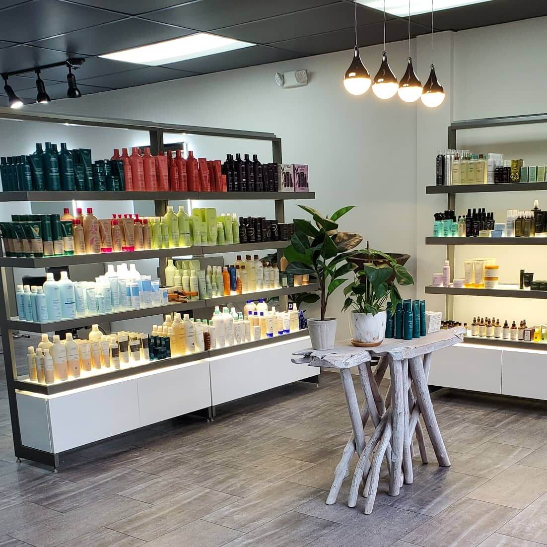 Shelves neatly display various hair and beauty products in a modern, well-lit salon, with potted plants and a small wooden table in the center.