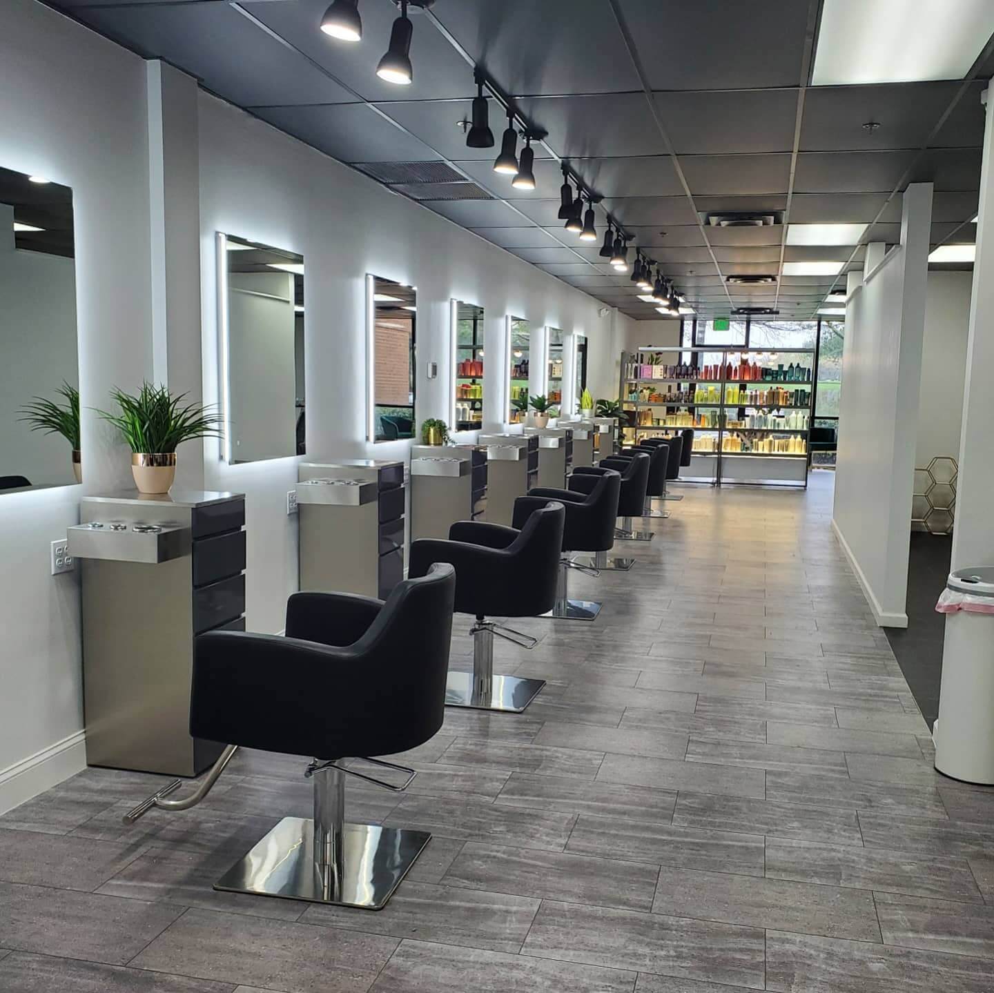 A modern hair salon with black styling chairs, illuminated mirrors, potted plants, and shelves of hair products in the background.