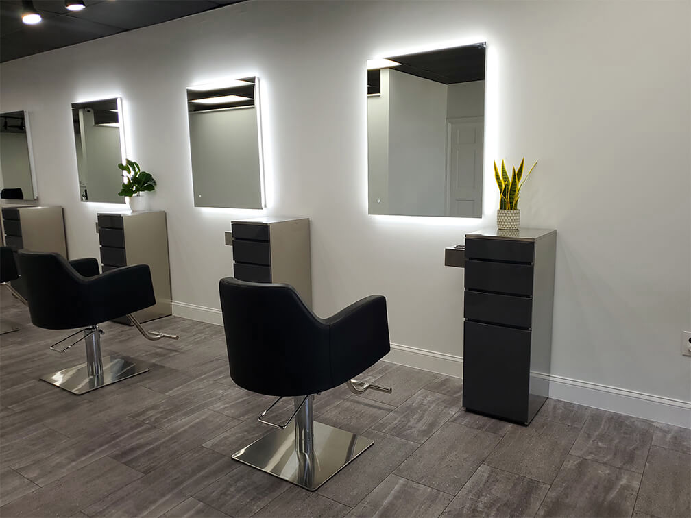 Modern salon interior with black styling chairs, illuminated wall mirrors, gray cabinets, and potted plants on top of the cabinets, set against a white wall and tiled floor.