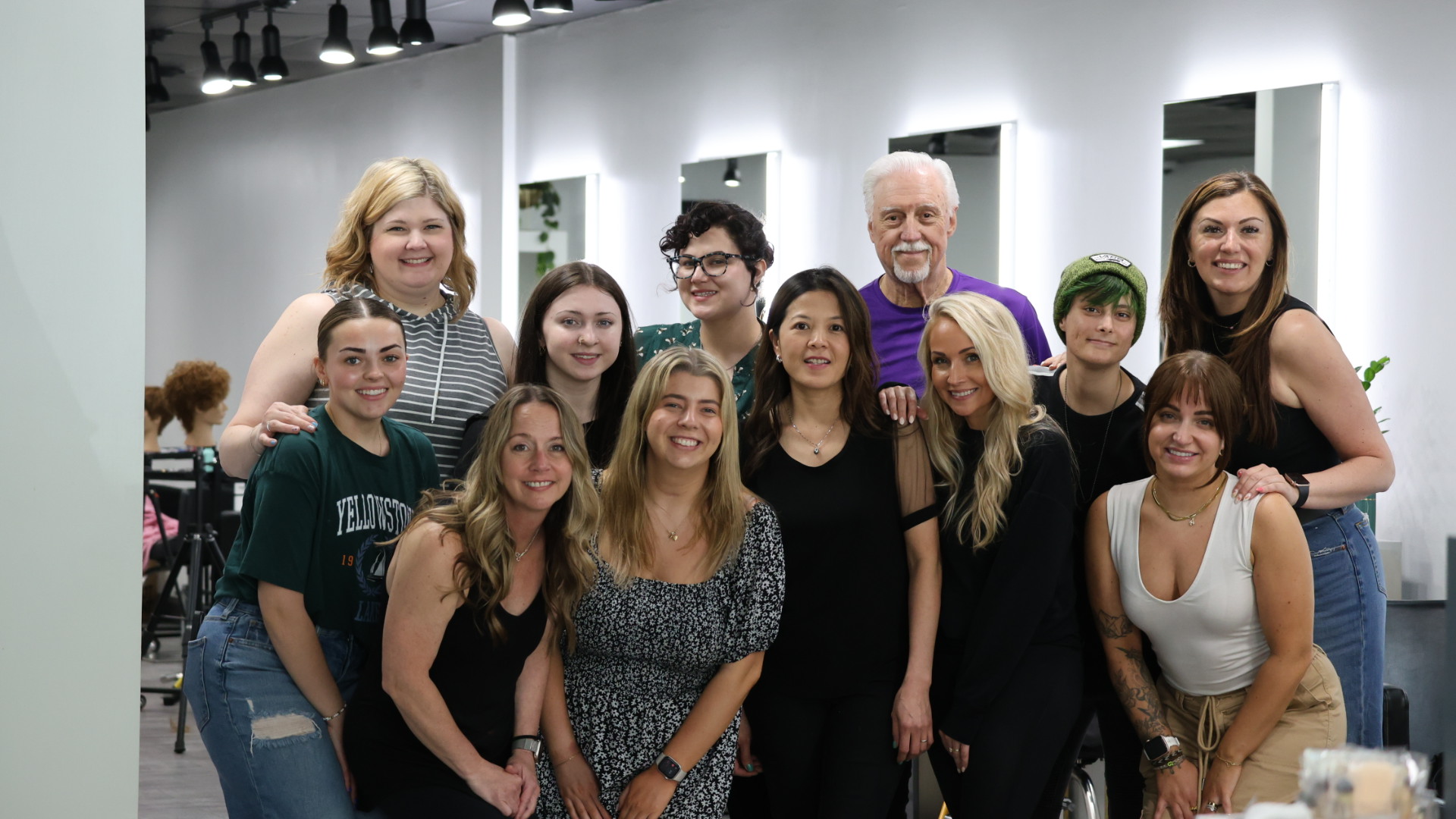 A group of eleven people pose indoors, all wearing black tops and blue jeans or pants, standing on a gray floor under a black ceiling.