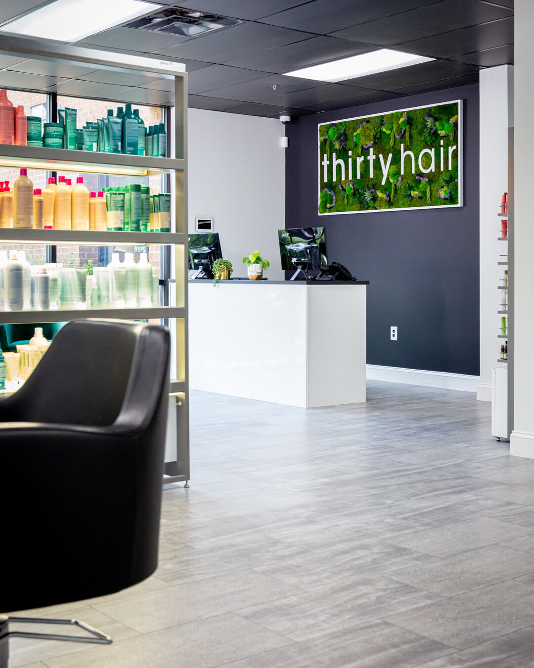 Modern hair salon interior with a black styling chair, shelves of hair products, and a reception desk with a "thirty hair" sign on the wall behind it.