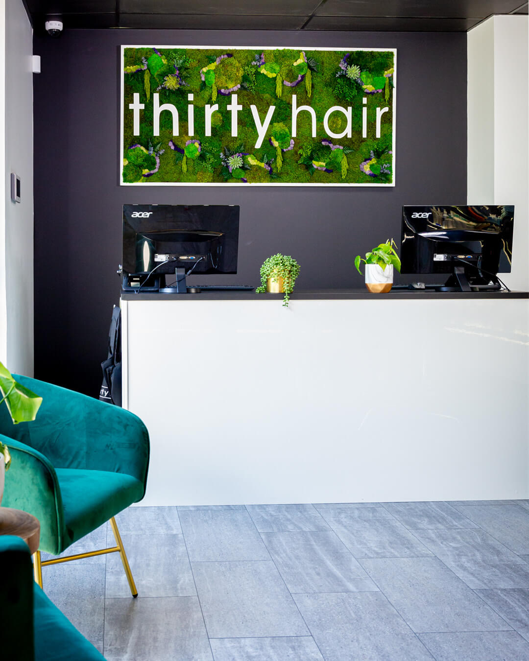 Reception desk with two computer monitors, potted plants, and a wall sign reading "thirty hair" above, in a modern salon interior.