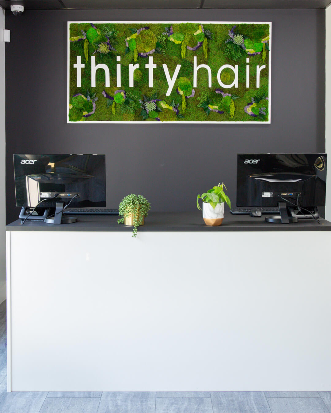 Reception desk with two computer monitors and potted plants, with a green moss wall art displaying the words "thirty hair" above.