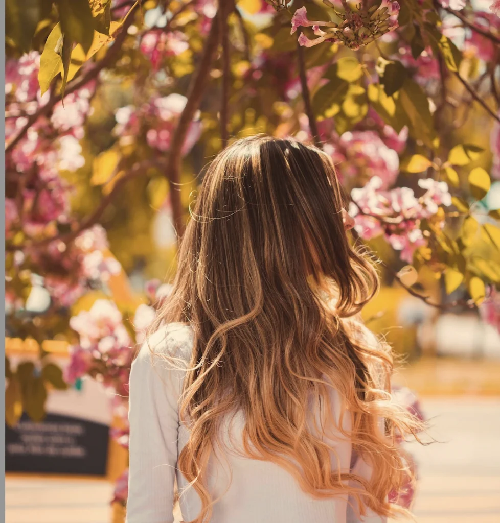 A person with long, wavy, blonde hair stands outdoors facing away, surrounded by blooming pink flowers and green leaves in bright sunlight.