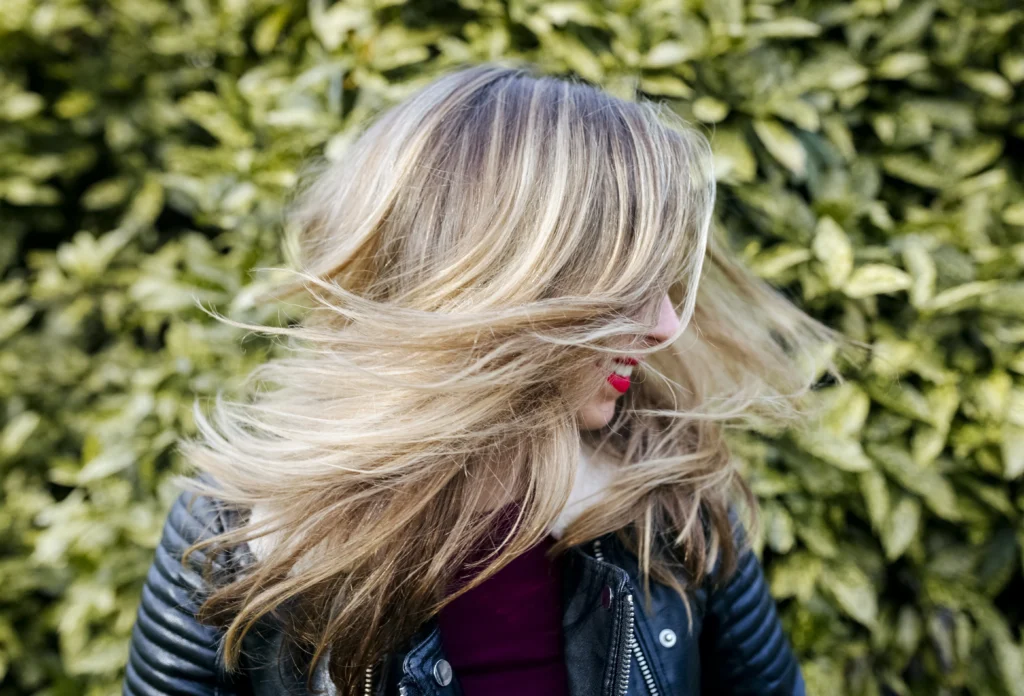 Person with long, blonde hair wearing a black leather jacket, turning their head in front of leafy green foliage.
